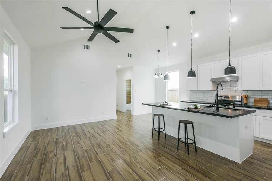 This is a spacious, open-concept kitchen with modern white cabinetry and a large central island featuring a cooktop and bar seating. The room is well-lit with natural light, pendant lights, and recessed lighting, complemented by a high vaulted ceiling with a ceiling fan. Elegant wood flooring runs throughout the space.