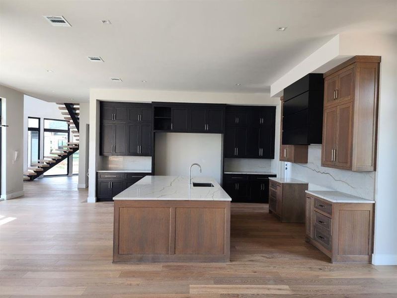 Kitchen with an island with sink, backsplash, light stone countertops, light wood-type flooring, and sink