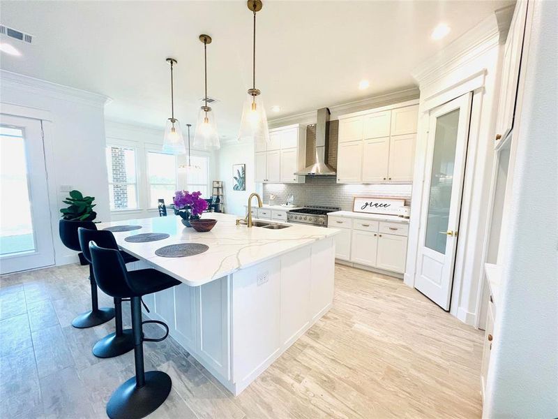 Kitchen with pendant lighting, wall chimney exhaust hood, double oven range, a center island with sink, and backsplash