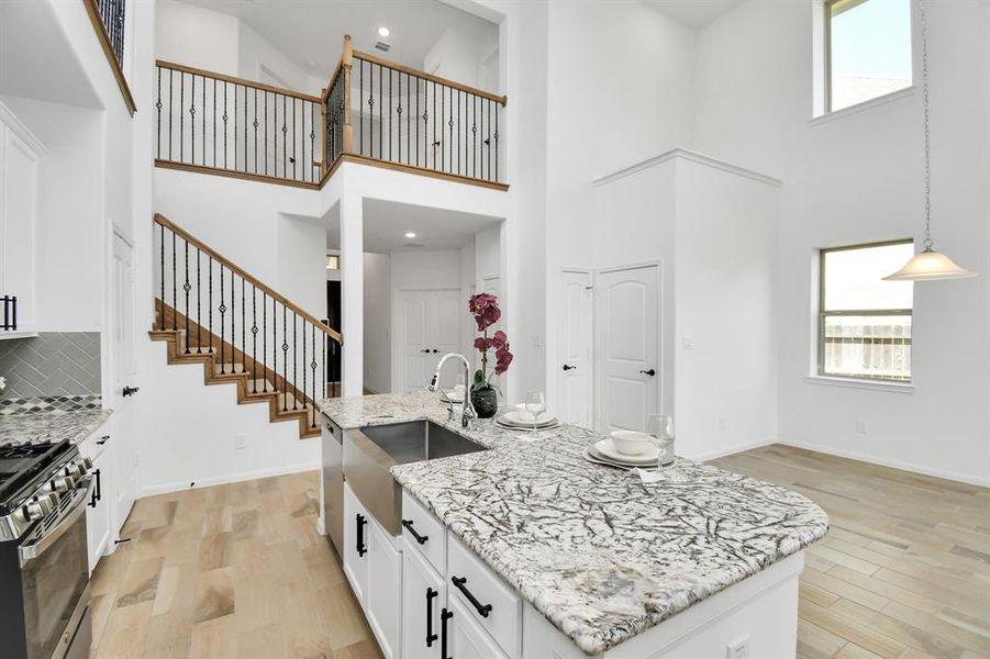 You're looking at a spacious, modern kitchen with high ceilings, featuring granite countertops, stainless steel appliances, and a stylish staircase leading to an upper level. The area is well-lit with natural light from windows and an elegant pendant light.