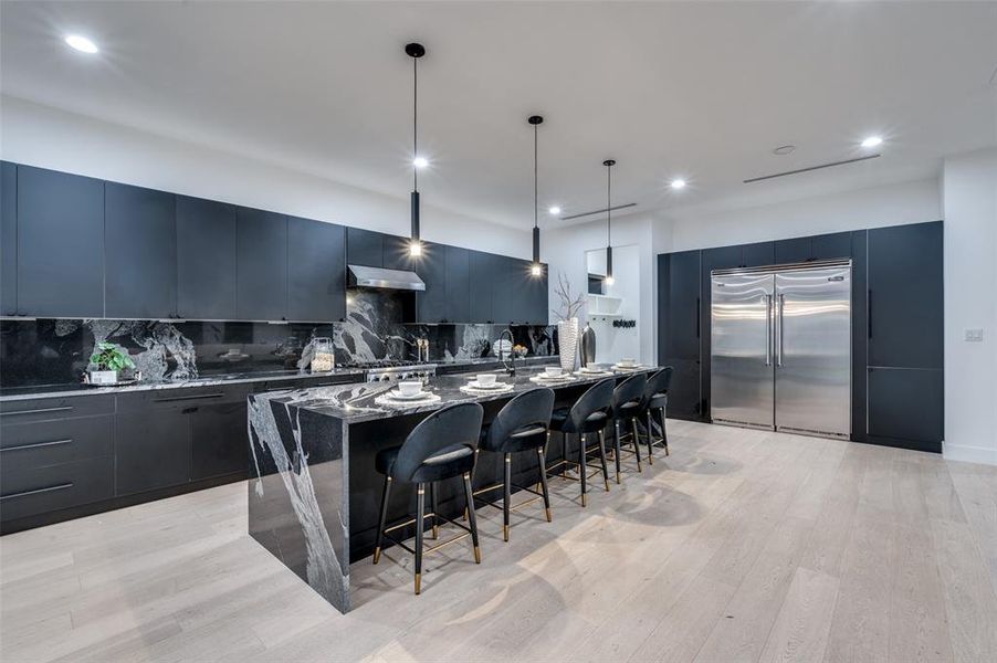 Kitchen with a large island with sink, stainless steel built in fridge, decorative backsplash, hanging light fixtures, and light hardwood / wood-style floors