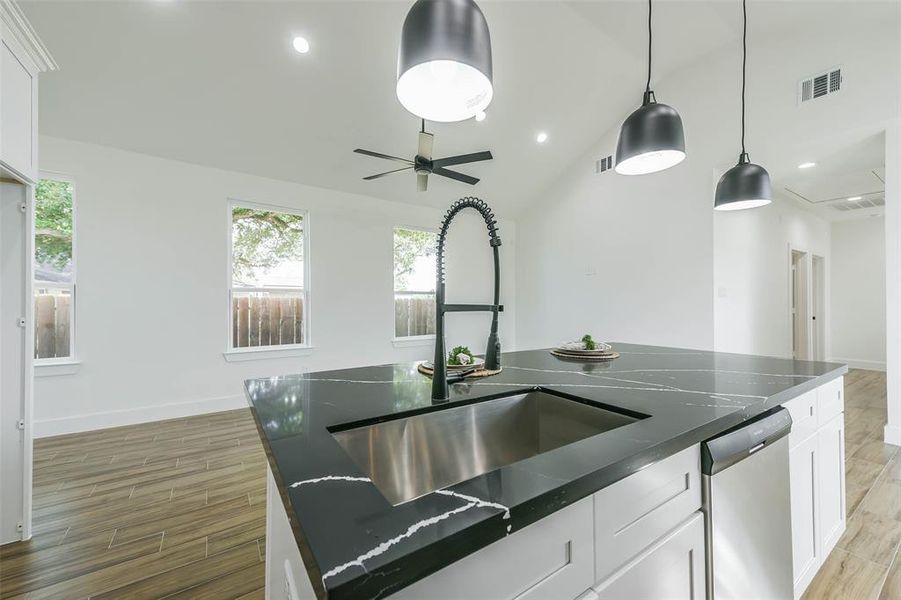 This is a modern, well-lit kitchen featuring a large island with a sleek countertop, stainless steel sink, contemporary pendant lighting, and hardwood flooring. The space has a clean, minimalist design with ample natural light from multiple windows and is complete with a ceiling fan and recessed lighting.