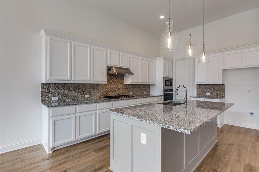 Kitchen with tasteful backsplash, light wood-type flooring, pendant lighting, white cabinets, and a kitchen island with sink