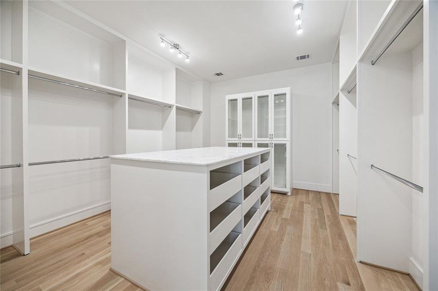 The ultimate dream closet for fashion enthusiasts! It boasts a central packing and folding island with drawer storage, multiple hanging racks, a built-in glass-front cabinet, sleek track lighting, and elegant wood flooring.