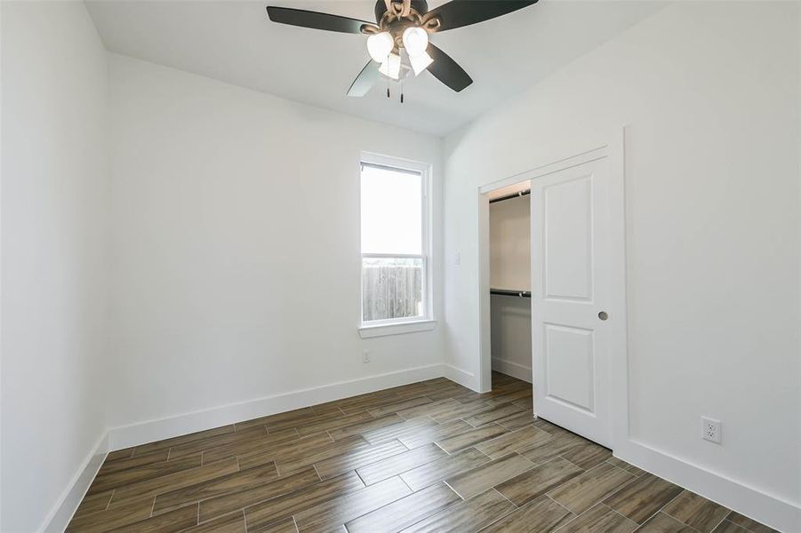 This is a bright, empty room with clean white walls, wood-patterned tile flooring, a ceiling fan with lights, and a window providing natural light. There's a closed closet door on the right.