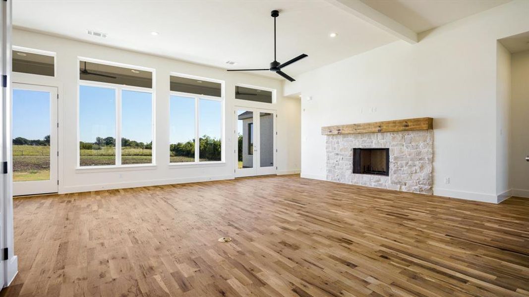 Unfurnished living room featuring a stone fireplace, beamed ceiling, light wood-type flooring, and ceiling fan