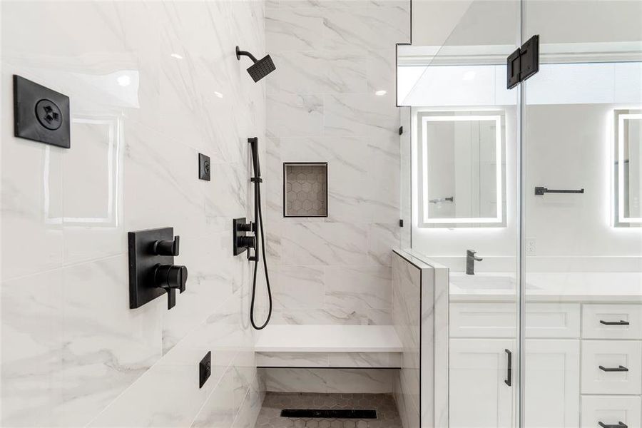 The bathroom features sleek, modern design with white marble walls, rain shower head, dual wall shower heads and body sprays, a built-in bench, and matte black fixtures. There's ample cabinetry for storage, and the vanity includes a well-lit mirror.