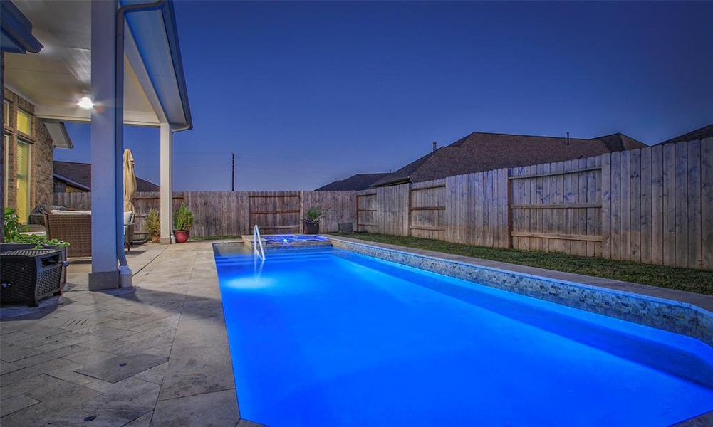 This photo features a beautifully lit backyard with an inviting in-ground pool, surrounded by a spacious patio area and privacy fence, perfect for relaxing or entertaining.