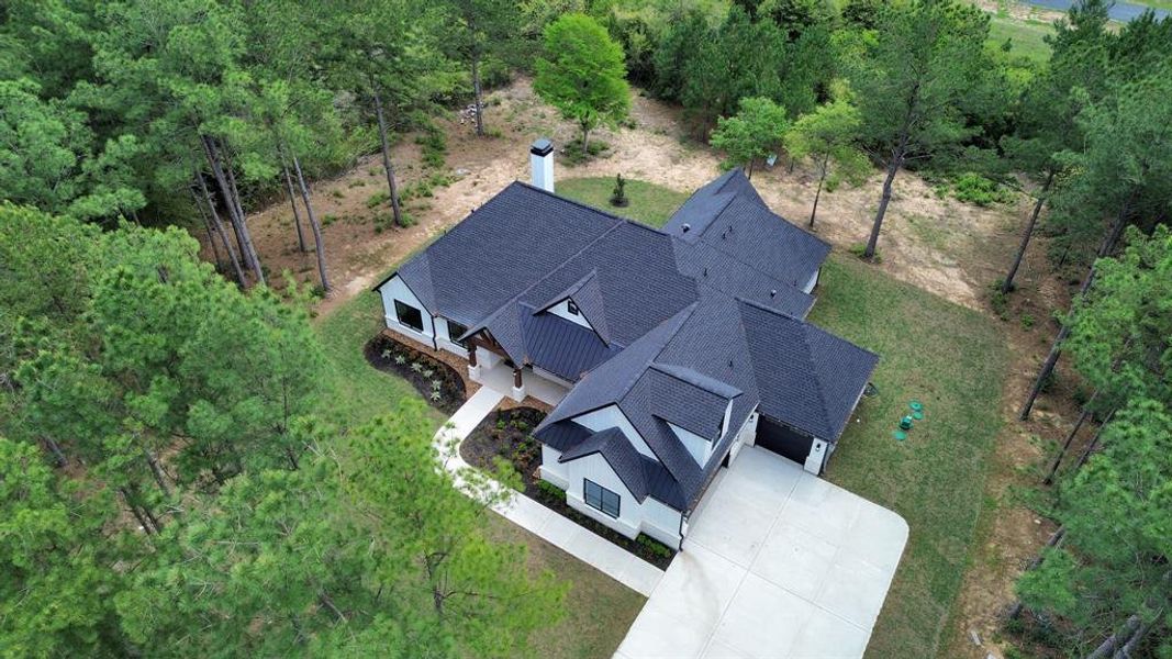 This is an aerial view of the home with a dark roof, surrounded by a lush pine forest, offering privacy and a close connection to nature. The property includes a well-maintained lawn and a large driveway.