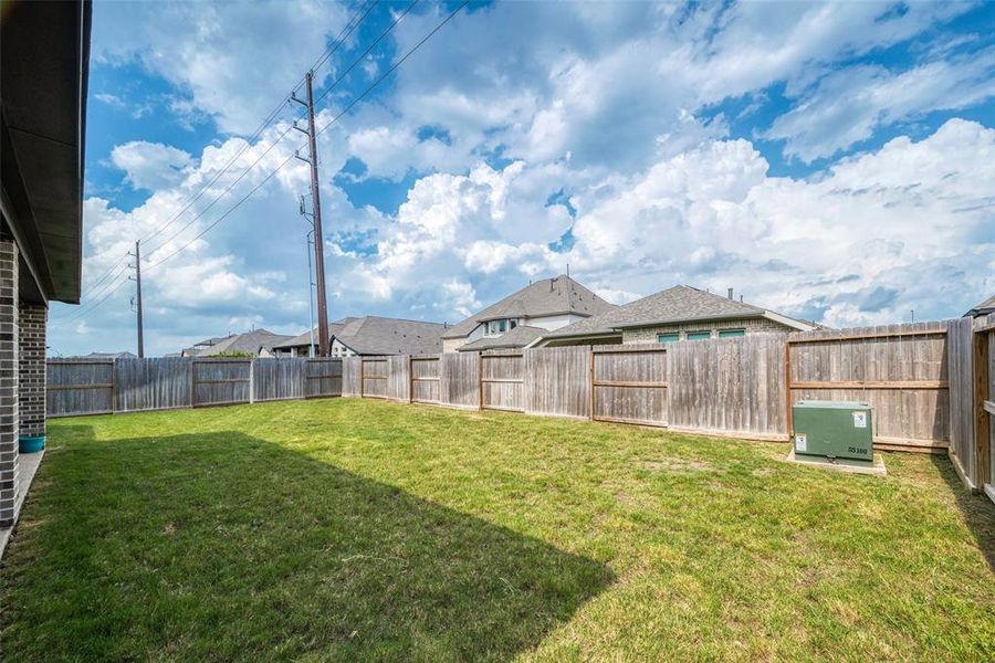 This is a spacious, grassy backyard with a wooden privacy fence, presenting clear skies above and the exterior of neighboring houses. A utility box sits near the fence.