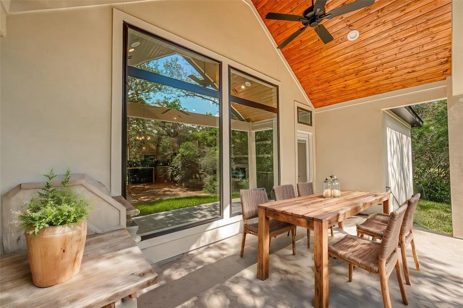 This is a spacious, covered outdoor patio featuring a wooden dining set, a warm-toned wood ceiling with a ceiling fan, and large windows providing a view of the lush backyard.