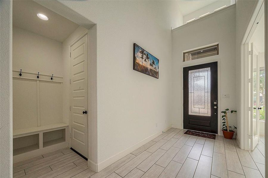 This is a bright entryway featuring high ceilings, a modern front door with glass panels, a built-in coat rack and bench, and tiled flooring. A welcoming space with ample natural light.