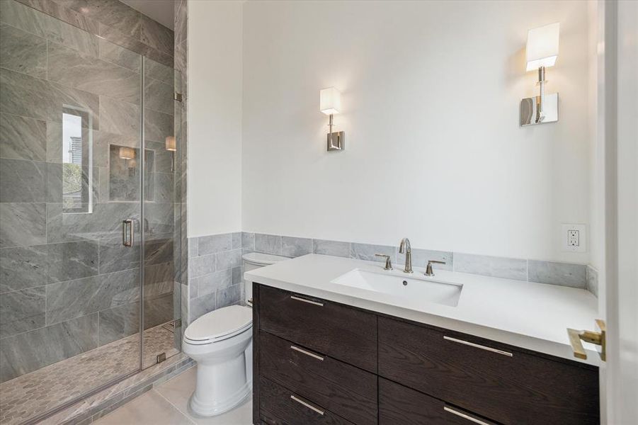 This luxurious ensuite bathroom showcases a walk-in shower with gorgeous marble floor and wall tile, creating a spa-like atmosphere. Quartz countertops, designer polished nickel sconces, and a striking ebonized oak vanity add to the space's refined elegance.