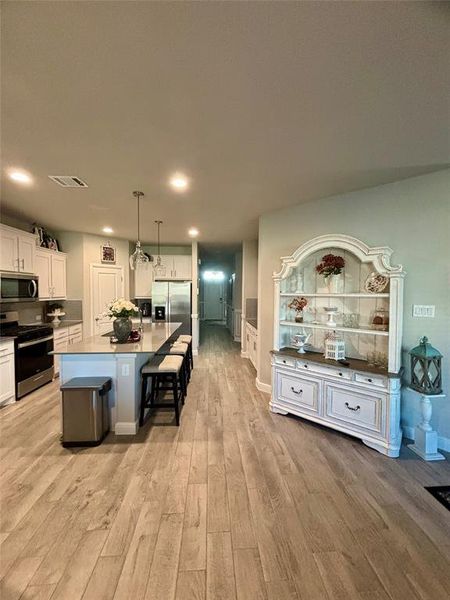 Kitchen featuring light hardwood / wood-style floors, stainless steel appliances, hanging light fixtures, white cabinets, and an island with sink