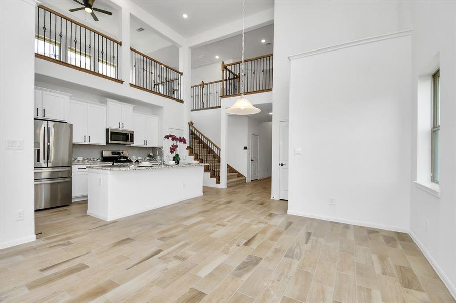 This is a spacious, modern open-plan living space with high ceilings, featuring a sleek kitchen with stainless steel appliances and a central island. The area is well-lit with natural light and includes an elegant staircase leading to an upper level with a balcony overlooking the living area.