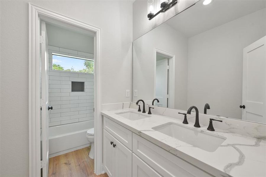 This is a modern, bright bathroom with dual sinks, quartz countertops, and white cabinetry. It features a bathtub with subway tiles and a window that brings in natural light.