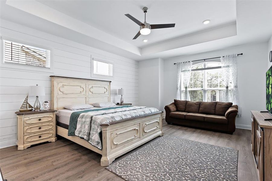 The primary suite is oh so spacious, featuring a large seating area, stepped up ceiling, ceiling fan amd high bed wall windows all surrounded by the shiplapped accent wall