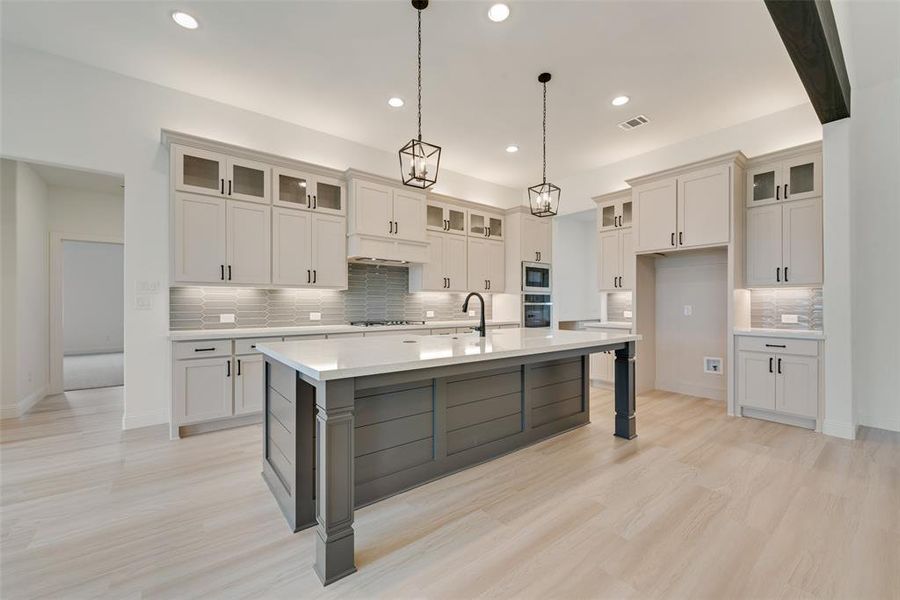 Kitchen with hanging light fixtures, backsplash, appliances with stainless steel finishes, light hardwood / wood-style floors, and an island with sink