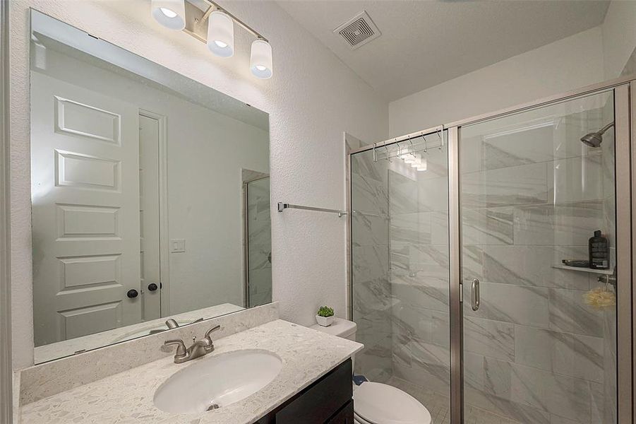This is a modern bathroom featuring a single sink vanity with a granite countertop, a large mirror, a glass-enclosed shower with gray marble-like tiles, and simple overhead lighting. The space is neat and well-lit, with a neutral color scheme.