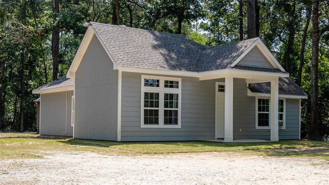 Home featuring a gabled roof design, neutral siding, and white trim around windows and the front door. The house is set against a backdrop of trees with a clear front yard space.