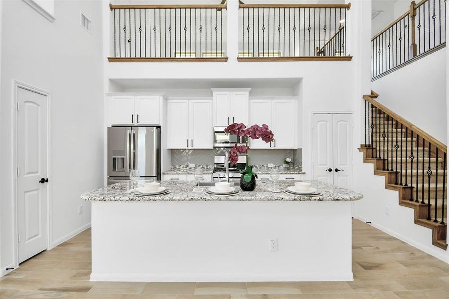 This is a spacious, modern kitchen with white cabinetry, granite countertops, and stainless steel appliances, featuring an island and elegant open staircase leading to the upper level.