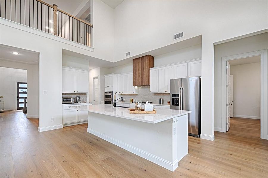 Kitchen with light hardwood / wood-style flooring, a high ceiling, white cabinets, a kitchen island with sink, and appliances with stainless steel finishes