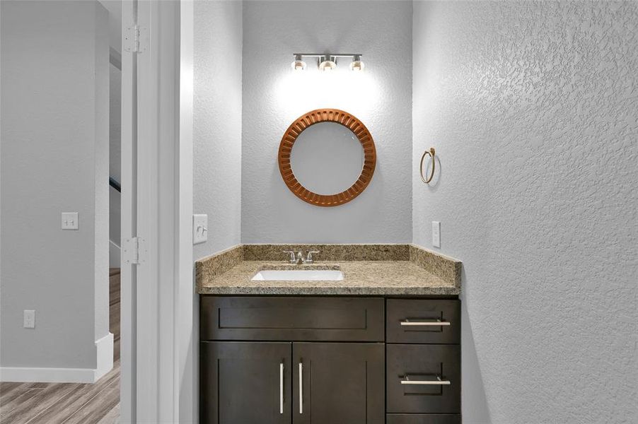 This is a modern half-bathroom with a granite countertop vanity, undermount sink, dark wood cabinetry, and a circular mirror.