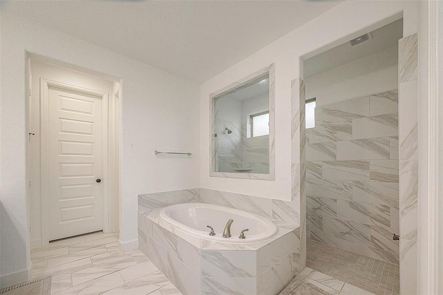 This is a modern bathroom featuring a large corner bathtub with marble-like tile surrounds, a separate walk-in shower area, sleek fixtures, and a spacious vanity mirror. The room has neutral colors offering a clean and serene atmosphere.