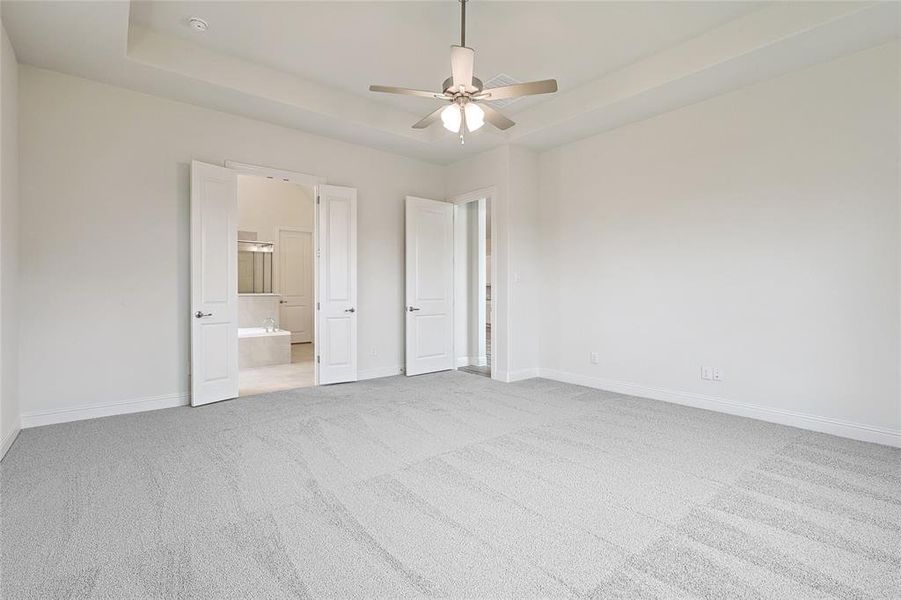 Unfurnished bedroom featuring ceiling fan, light colored carpet, a tray ceiling, and ensuite bath