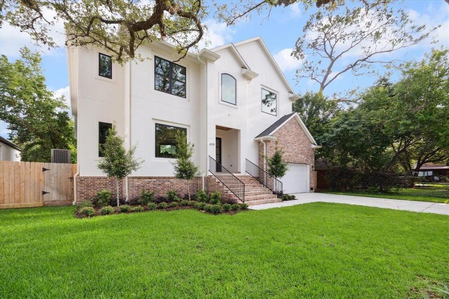 You're looking at a modern two-story home with a fresh white facade and brick accents, featuring large windows and an inviting entrance with stairs. The property includes a well-manicured lawn and mature trees, with a private driveway leading to an attached garage.