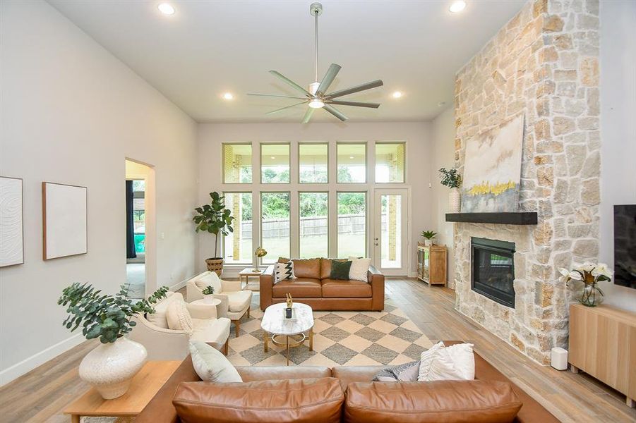 This is a spacious, open-concept living room with high ceilings and a modern ceiling fan. It features a cozy stone fireplace, a seamless transition into the dining and kitchen areas. The room features natural light throughout.