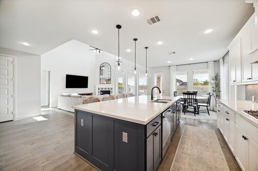 This is a modern open-plan living space featuring a spacious kitchen with a large island and white cabinetry, leading directly into a cozy living area with a mounted TV and fireplace. The room is well-lit with natural light from large windows and stylish pendant lighting.