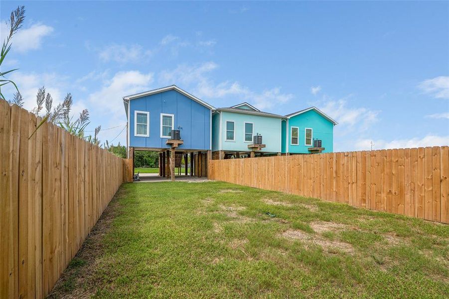 The photo shows back of the home with a vibrant blue exterior, elevated on stilts, featuring a spacious fenced backyard with ample green space.