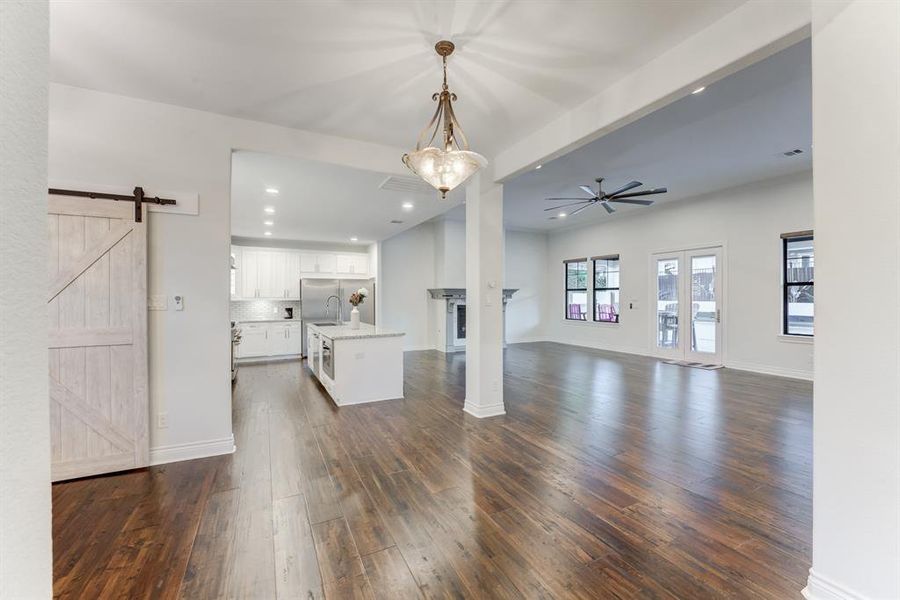 A spacious, open-concept living area, featuring a modern kitchen with white cabinetry and a central island. The room is well-lit with natural light from multiple windows and a glass door leading outside. A rustic sliding barn door adds a charming touch to the space.