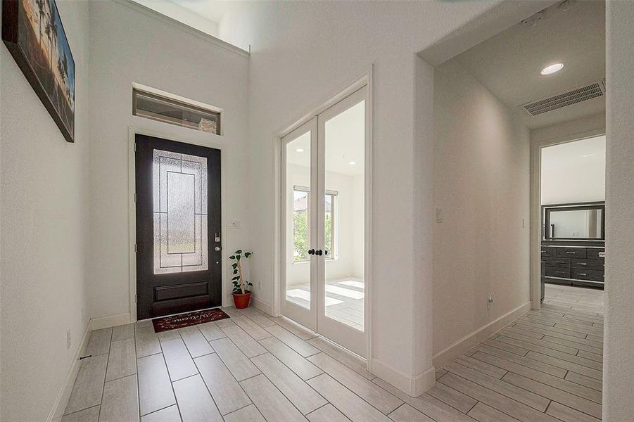 This is a bright and welcoming entryway featuring a high ceiling, tiled flooring, and a modern front door with glass details, leading to a well-lit corridor with access to other rooms.