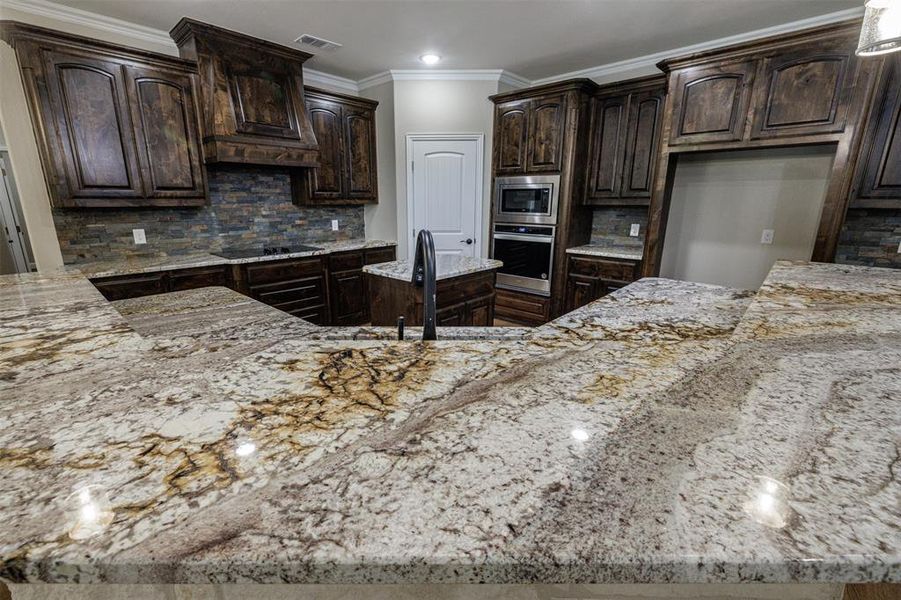 Kitchen with light stone counters, ornamental molding, dark brown cabinets, backsplash, and stainless steel appliances