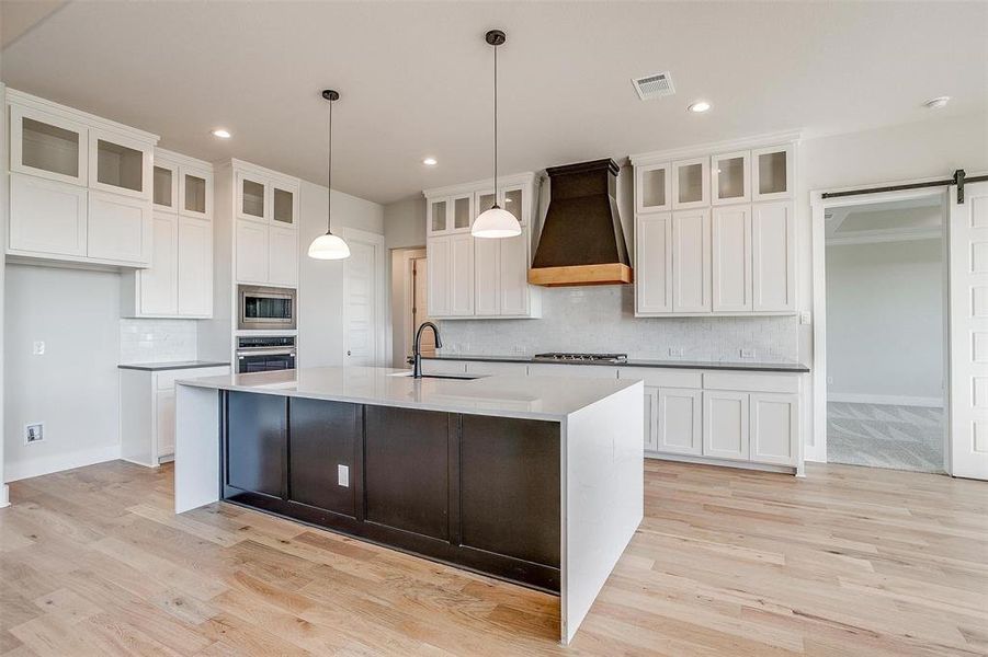 Kitchen featuring custom exhaust hood, light wood-type flooring, a barn door, a center island with sink, and appliances with stainless steel finishes