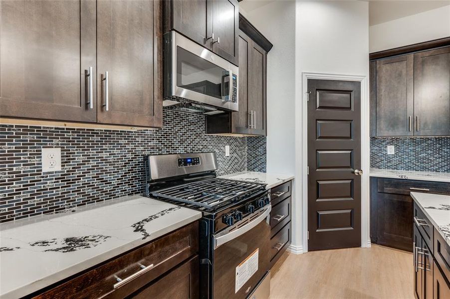 Kitchen with light stone countertops, dark brown cabinets, light hardwood / wood-style floors, stainless steel appliances, and decorative backsplash