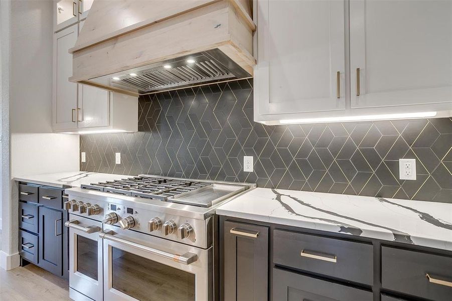 Kitchen with high end stainless steel range oven, tasteful backsplash, light stone countertops, light wood-type flooring, and premium range hood