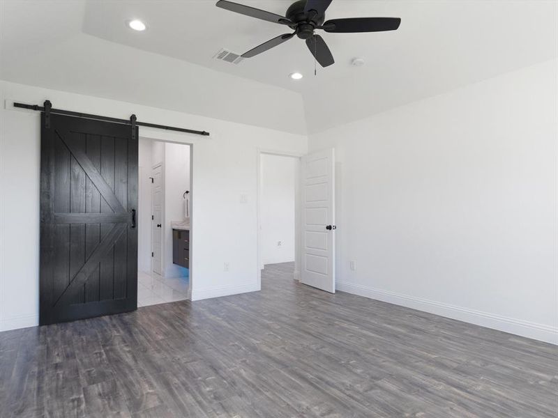 Unfurnished bedroom featuring ensuite bathroom, a barn door, wood-type flooring, and ceiling fan