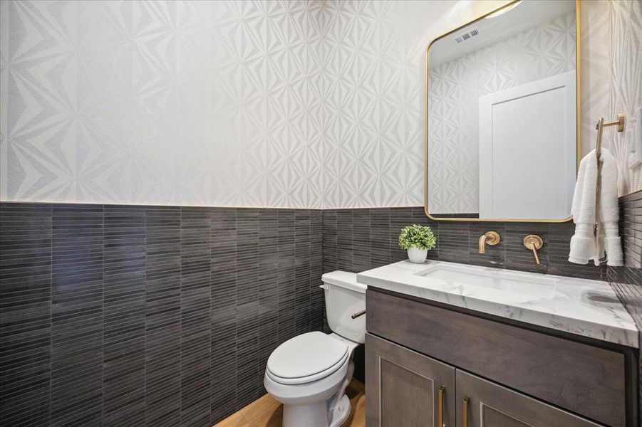 Designer powder bath features custom wallpaper and tiled walls, wall-mounted plumbing fixtures, and brushed gold hardware.