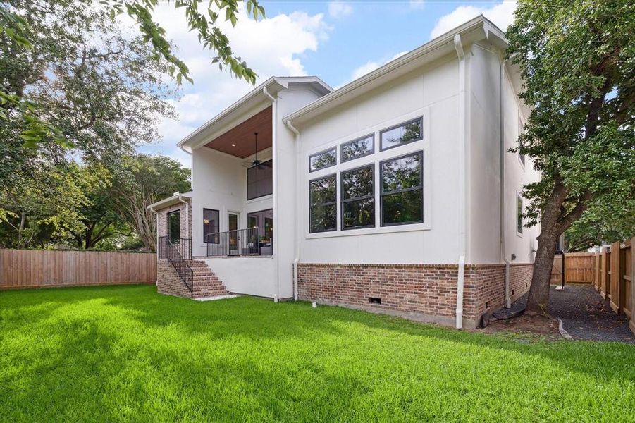 This is a modern two-story home featuring large windows, an elevated covered patio with a wooden ceiling, and a spacious, well-maintained lawn. The property is fenced for privacy and includes mature trees.
