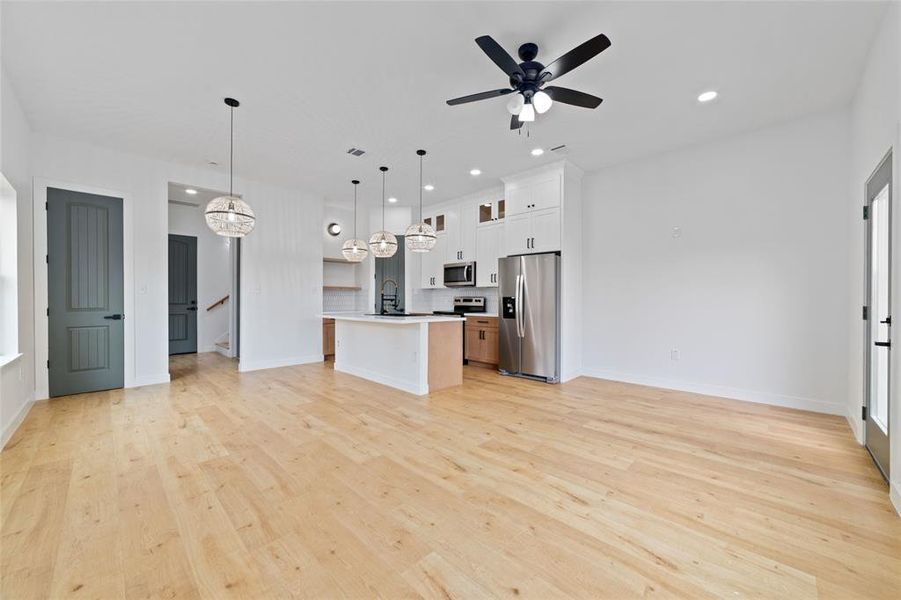 This modern, open-concept living space features a bright kitchen with stainless steel appliances and an large island, complemented by elegant light fixtures and vinyl plank wood flooring throughout. The area is well-lit with natural light from windows and enhanced by recessed lighting and ceiling fans.