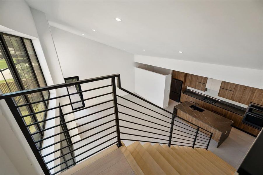 This is a modern, open-plan interior with a high ceiling, featuring a sleek kitchen with wood finishes and built-in appliances. The view from the top of a wooden staircase with a stylish metal railing overlooks the space, highlighting the bright and airy feel of the home.