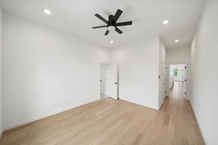 This is a bright, modern room with clean lines, featuring light hardwood floors, white walls, and a black ceiling fan. There's a closet with white doors and recessed lighting, leading to a hallway that suggests a seamless flow throughout the home.