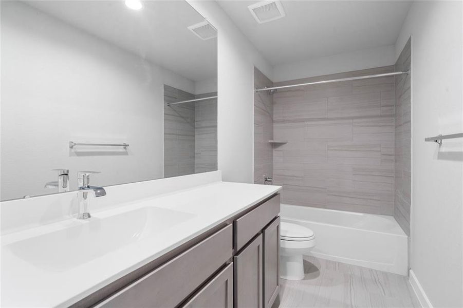 Secondary bath features tile flooring, bath/shower combo with tile surround, dark stained cabinets, beautiful light countertops, mirror, dark, sleek fixtures and modern finishes.