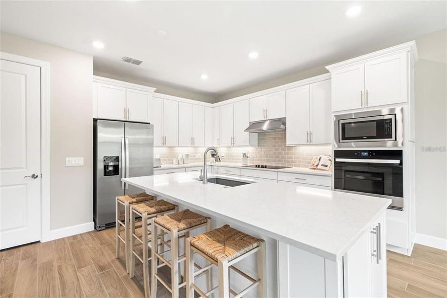 It features a large island, walk-in pantry, quartz countertops, upgraded 42-inch wood shaker-style cabinetry, composite single-bowl sink, cooktop with stainless steel hood, stainless steel fridge, subway tile backsplash, and built-in microwave and oven.