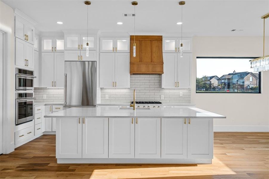 Kitchen with custom exhaust hood, hanging light fixtures, appliances with stainless steel finishes, an island with sink, and white cabinetry