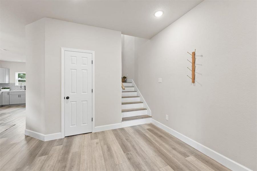 This is a modern, freshly painted space with a view into a bright kitchen, featuring wood-look flooring and white walls. A staircase leads upstairs, and there's a wooden coat rack on the wall.