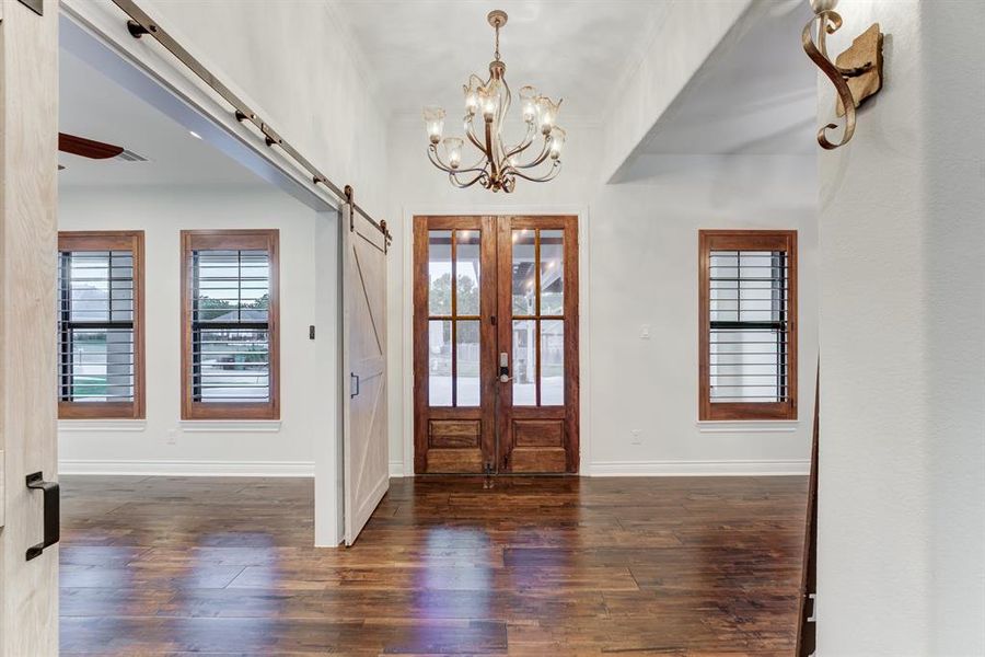 A bright, spacious entryway features rich hardwood floors and elegant double doors with glass panels, providing ample natural light. A classic chandelier and a modern sliding barn door add a mix of traditional and contemporary touches to the space.